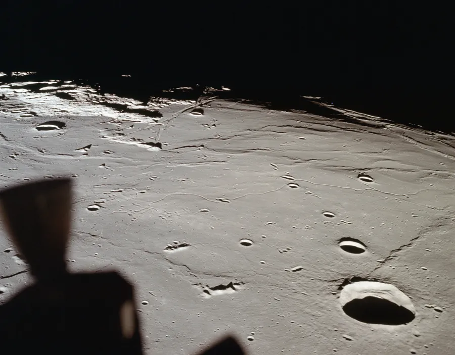 A grey lunar plain under low sunlight, scattered with craters and thin rilles, curving to a dark horizon. A thruster of the Lunar Module is silhouetted in the corner.