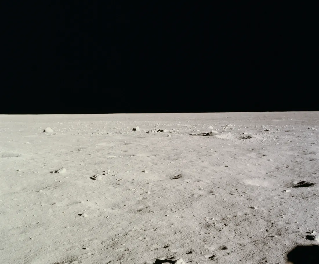 A flat grey lunar plain scattered with small rocks, under a black sky, seen from the lunar module window.