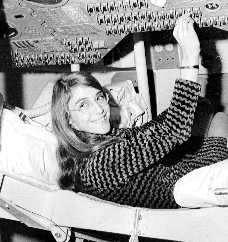 Margaret Hamilton, smiling over her shoulder, reclines in a command-module seat and reaches up to the switch panel.